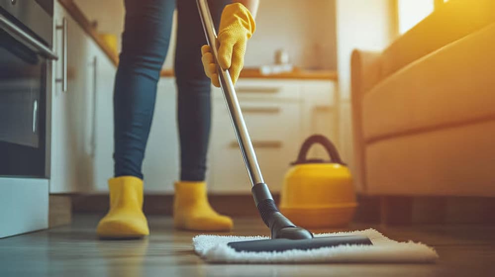 Person in yellow gloves mopping a kitchen floor with a steam cleaner and yellow vacuum nearby.